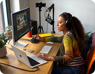 امرأة في منزلها تعمل خلف مكتبها على MacBook Pro متصل بشاشتين خارجيتين وترتدي سماعات رأس وتتحدث في الميكروفون