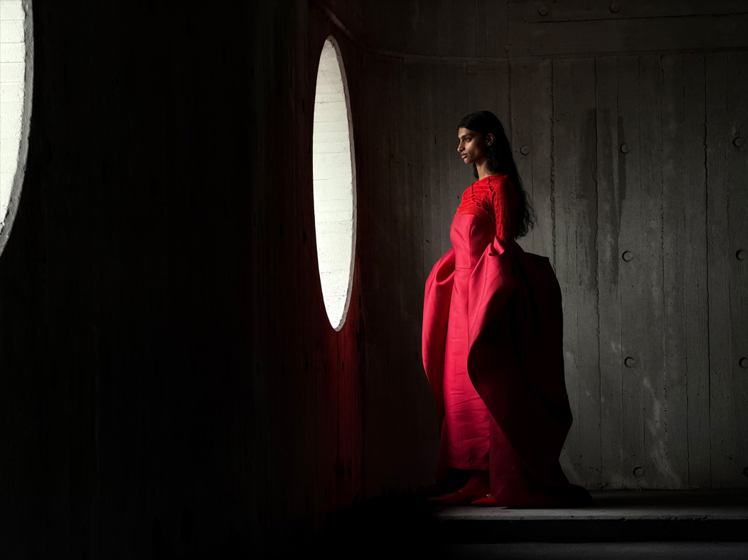 Une femme vêtue d’une longue robe rouge se fait photographier dans une pièce vide. L’app Appareil photo change de distance focale pour illustrer les capacités du système caméra pro, mettant en valeur : 100 mm